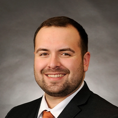 Chiropractor Andrew Holzli In Suit Smiling In Front OfGrey Background